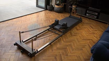 A Pilates reformer machine with a black padded carriage and metal frame, placed on a polished herringbone wood floor in a modern living room setting.